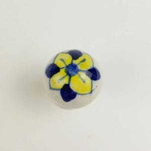Può includere: Un piccolo pomello rotondo in ceramica con un motivo floreale dipinto a mano. Il fiore presenta petali gialli, dettagli blu scuro e un centro blu. Il pomello è bianco sporco e probabilmente utilizzato per mobili o scopi decorativi.