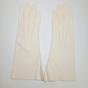 May include: A pair of off-white leather gloves featuring three vertical stitched lines on the back of each hand. The gloves extend past the wrist and have a smooth, matte finish with clean edges and a simple, unadorned design throughout.
