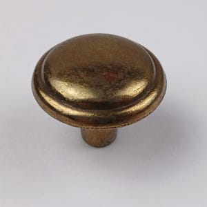 Könnte beinhalten: Ein bronzefarbener Schrankknauf mit abgerundeter Oberseite und kurzem Stiel. Der Knauf hat eine leicht strukturierte Oberfläche und einen dekorativen Rand. Geeignet für Schubladen und Schränke.