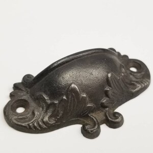 May include: A dark gray, cast metal cup pull handle featuring a curved, semi-circular grip and decorative, ornate leaf-like detailing on the mounting plates. The hardware includes two circular screw holes on the ends for secure attachment to furniture.