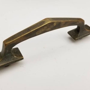 Puede incluir: Un tirador de armario de latón con un diseño geométrico. El tirador tiene una forma rectangular con una parte superior curva y dos placas cuadradas en cada extremo.