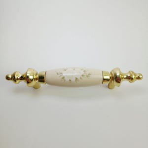 May include: A drawer pull handle featuring a central cream-colored ceramic cylinder with a subtle white floral pattern. The handle is flanked by two polished gold-tone metal end pieces with a tiered, rounded shape on each side of the center.