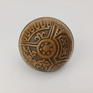 May include: A bronze-colored, antique door knob with intricate, embossed floral and geometric patterns. The round knob features a central circular design surrounded by detailed embellishments, showcasing a vintage aesthetic.