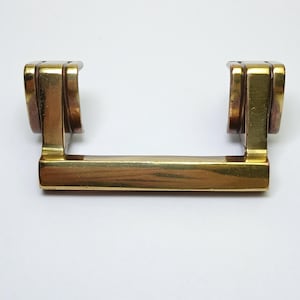 May include: A polished brass drawer pull featuring a horizontal rectangular bar handle connected to two vertical, curved mounting brackets. The metal surface has a reflective, gold-toned finish with subtle variations in color and texture.