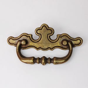 Op de afbeelding: Antieke messing ladeknop met een sierlijk ontwerp. De knop heeft een gebogen handvat en een gedetailleerde achterplaat met een centraal bloemmotief. Deze hardware is waarschijnlijk voor een kast of dressoir.