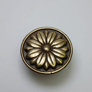 Puede incluir: Un pomo de mueble redondo de metal color latón antiguo con un diseño floral simétrico en relieve, con doce pétalos que irradian desde un punto central circular. El acabado presenta una textura metálica ligeramente desgastada.