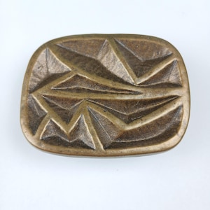 以下が含まれることがあります： 幾何学的な抽象的なデザインが施された長方形のブロンズ色のトレイ。トレイはテクスチャ加工された表面とわずかに盛り上がった縁を持っています。