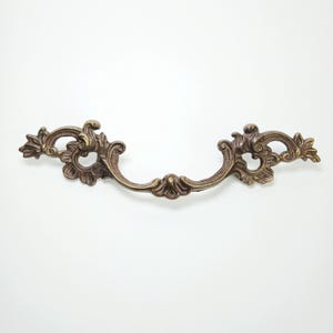 Peut inclure: Une poignée de tiroir en métal de couleur bronze présentant un design orné et symétrique avec des motifs de feuilles en volutes et une poignée centrale incurvée. La quincaillerie a une finition antique patinée avec des zones sombres.