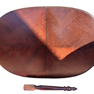 May include: Oval wooden table with a rich, dark brown finish. The tabletop is divided into two sections, with visible wood grain and a glossy sheen. A detached, matching wooden table leg is also visible.