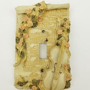 May include: Decorative light switch cover with a musical theme. The cover is beige with a scroll design, musical notes, a violin, and pink roses. The cover has a single light switch opening and two screw holes.