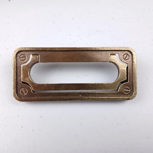 Puede incluir: Un tirador de cajón rectangular de latón con una abertura rectangular en el centro. El tirador tiene un acabado ligeramente envejecido, antiguo.