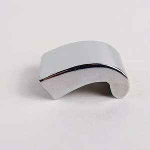 May include: A small, curved, chrome-finished metal cabinet pull or handle component. The object features a smooth, reflective, silver-colored surface with a rounded top edge and a hollowed-out underside, designed for attachment to a flat surface.