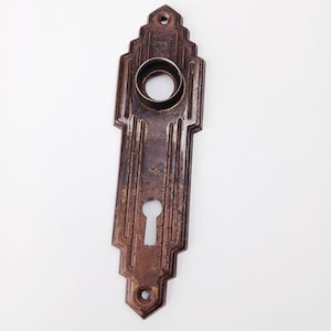 Può includere: Una placca per porta antica in stile Art Déco con una serratura e un'apertura circolare per una maniglia. La placca metallica ha una finitura scura e invecchiata con scanalature verticali e dettagli decorativi. Placca rettangolare con parte superiore decorativa.