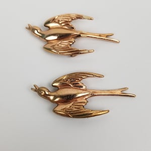 May include: Two gold-toned metal bird brooches in the shape of a swallow. The birds are in flight with their wings spread out.