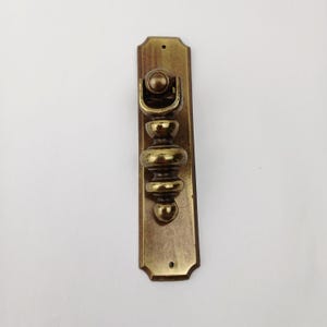 May include: A vintage brass door pull with an ornate design. The rectangular backplate has a decorative top and bottom edge. The pull features a series of rounded and bulbous shapes, with a curved handle at the top. The hardware is a warm, antique gold color.