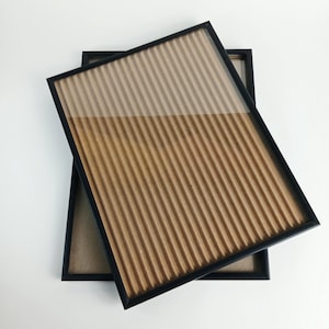 May include: Two rectangular picture frames with thin black borders. The top frame is angled over the bottom frame, both containing a textured, light brown corrugated cardboard insert that fills the entire display area behind the clear glass.