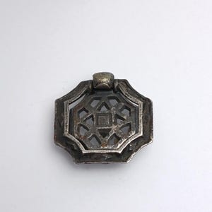May include: A tarnished silver-toned metal drawer pull featuring an octagonal frame with a central geometric lattice cutout design. The hardware includes a hinged, rounded bail at the top, showing signs of age and oxidation on the metallic surface.