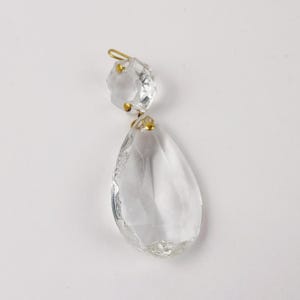 May include: A clear crystal chandelier pendant with a faceted top and a teardrop-shaped bottom. The pendant is connected with a gold-colored metal wire and small gold-colored metal accents. The crystal is transparent and reflects light.