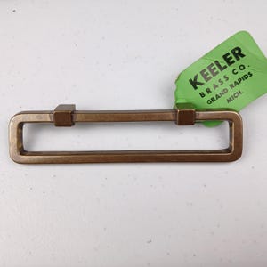 May include: A bronze cabinet pull with a rectangular shape and squared ends. The pull has a label attached that reads "Keeler Brass Co. Grand Rapids Mich."