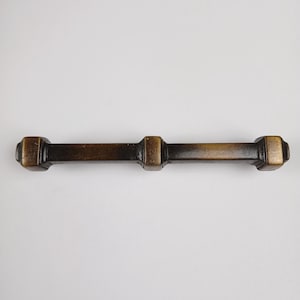 May include: A brass cabinet pull with a rectangular shape and a dark brown finish. The pull has three raised squares along its length.