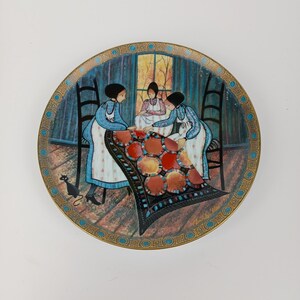 May include: Decorative plate featuring a folk art scene of three women quilting. The plate has a gold rim with a blue and gold border. The women are dressed in blue and white dresses, and a black cat is in the foreground.