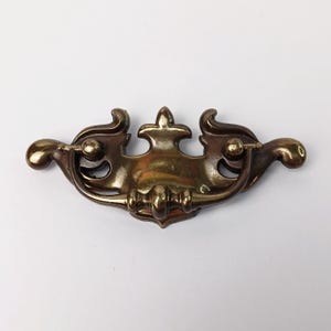 May include: Antique brass drawer pull with a decorative design. The pull features a central fleur-de-lis element flanked by curved, scroll-like handles. The hardware is approximately 6 inches long.