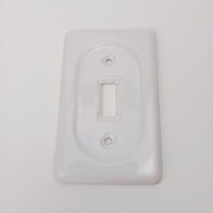 Pode incluir: Uma tampa de interruptor de luz branca, retangular, com uma reentrância oval em torno da abertura do interruptor. Possui dois orifícios para parafusos e um acabamento brilhante.