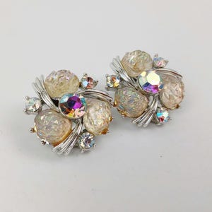 May include: A pair of vintage clip-on earrings. Each earring features a cluster of iridescent, clear, and peach-colored stones, accented with smaller, colorful crystals. The stones are set on a silver-toned metal base, creating a glamorous, eye-catching design.
