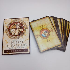 May include: A deck of Animal Dreaming Oracle Cards and a matching guidebook by Scott Alexander King. The cards feature a yellow background with a central compass-like emblem, while the guidebook cover displays animal illustrations in a circle.
