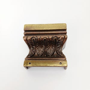 May include: A decorative furniture foot with a bronze finish. The foot features a detailed leaf design in a dark brown color, with bronze accents on the top and bottom. Two holes are visible on the bottom edge. This item is likely a component of a larger piece of furniture.