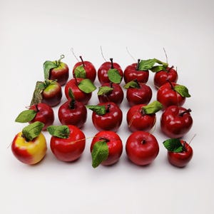 May include: A collection of artificial apples with a glossy red finish and green leaves. Some apples have a yellow and red gradient. The apples are arranged on a white surface, showcasing their realistic appearance.