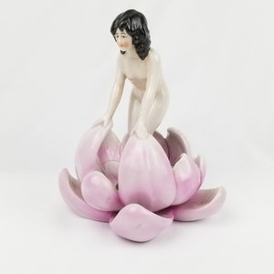 May include: A porcelain figurine of a woman with black hair, sitting in a pink lotus flower.