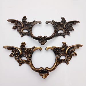 May include: Two matching antique brass-toned metal drawer pulls featuring an ornate, symmetrical scrollwork design with wing-like extensions on each side. The hardware has a dark, weathered finish with highlighted edges and a central cutout.