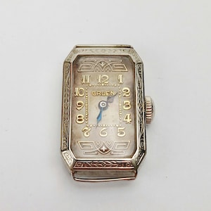 May include: A vintage Gruen wristwatch with a rectangular silver-toned face. The watch features gold-coloured numerals and hands, with the brand name "Gruen" displayed. The watch has an ornate, engraved bezel and a textured crown.