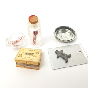 May include: A set of miniature baking supplies including a small glass jar with a cork stopper, a small metal cookie cutter in the shape of a person, a small metal baking sheet, and a small box with the text "Whitman's Sampler" printed on the top.