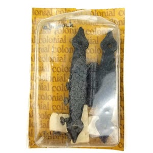 Op de afbeelding: Zwart decoratief scharnier in een doorzichtige plastic verpakking met het woord "colonial" gedrukt op de verpakking. Het scharnier heeft een textuur en een decoratief ontwerp. De verpakking is geel en wit.