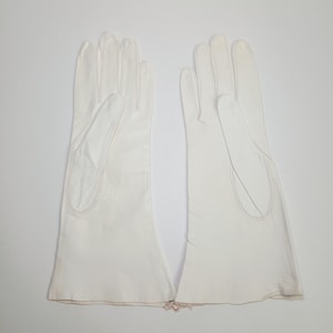 May include: A pair of white leather gloves featuring a smooth finish, four fingers, and a thumb section. The gloves extend past the wrist with a flared opening at the base. The stitching is visible along the seams of the fingers and the thumb.