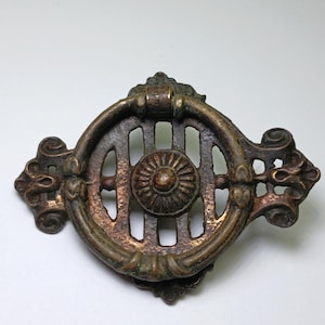 May include: An antique brass drawer pull featuring a circular ring handle attached to a decorative backplate. The backplate has a central rosette surrounded by vertical slats and ornate scrollwork on the left and right sides, with a weathered finish.