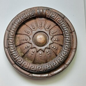 May include: A circular, dark brown metal door knocker featuring a central raised dome surrounded by a sunburst pattern of small dots. The outer ring is decorated with a repeating Greek key geometric motif, and a hinged pull ring is attached.