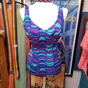 May include: A vintage-style, sleeveless top in shades of blue and purple, featuring a wavy pattern. The top is displayed on a mannequin. A rainbow-striped belt hangs nearby, and a vintage poster is in the background.