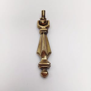 May include: A vertical brass furniture pull with a metallic gold finish. The hardware features a top mounting post, a hinged bail section, and a long, tapered, faceted body that ends in a small, rounded finial at the bottom of the piece.