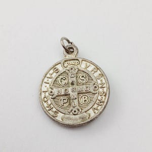 May include: A silver-colored pendant with a circular design. The pendant features a cross in the center, surrounded by text and symbols. The top of the pendant has a loop for attaching to a chain. The pendant is likely a religious item.