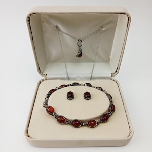 Könnte beinhalten: Ein Schmuckset in einer cremefarbenen Box. Das Set beinhaltet eine Halskette mit Anhänger, ein Armband und Ohrringe. Der Schmuck hat ovale, rotbraune Steine in einem silberfarbenen Metall.