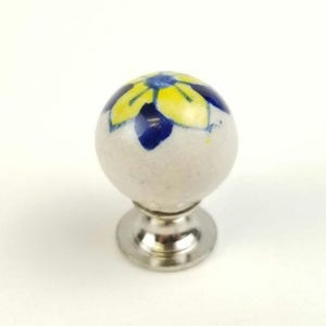 May include: A ceramic cabinet knob with a white sphere decorated with a yellow and blue floral pattern. The knob sits on a silver-coloured metal base, suitable for adding a decorative touch to furniture.