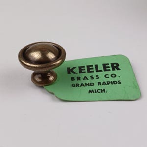 Könnte beinhalten: Ein bronzefarbener Schrankknopf mit abgerundeter Oberseite und Sockel. Der Knopf befindet sich neben einem grünen Etikett mit dem Text "KEELER BRASS CO. GRAND RAPIDS MICH."