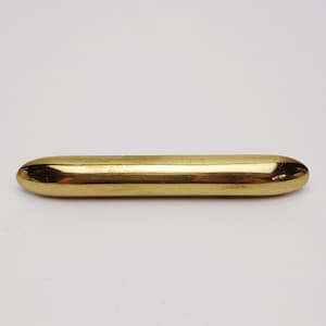 May include: A polished gold-toned metal bar with a smooth, cylindrical shape and rounded ends. The object features a reflective, metallic surface that catches the light, presenting a simple, elongated form with no additional markings or attachments.