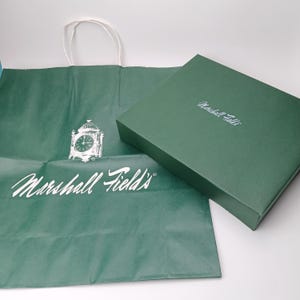 Op de afbeelding: Een groene papieren boodschappentas en een bijpassende doos. De tas heeft een witte klokafbeelding en de woorden "Marshall Field's" in witte letters. De doos heeft dezelfde tekst in een kleiner lettertype.