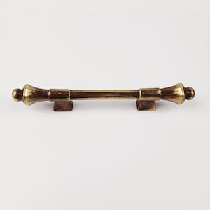 Peut inclure: Une poignée de meuble de couleur bronze avec une barre centrale et des extrémités décoratives. La poignée est fixée sur deux bases carrées, et le design a une esthétique vintage. La quincaillerie est sur fond blanc.