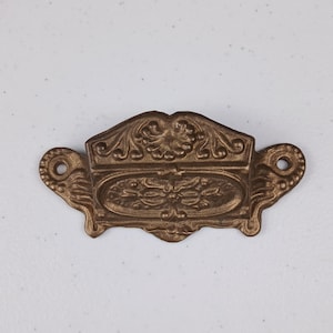 May include: A gold-toned metal drawer pull with an ornate design. The pull has a rectangular shape with a raised oval in the center. The edges of the pull are decorated with intricate floral patterns.