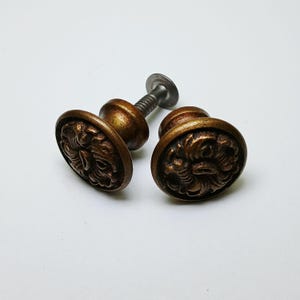 May include: Two round cabinet knobs with a dark bronze finish and a raised, intricate floral or scrollwork pattern on the face. Each knob features a central circular base and a threaded metal screw extending from the back for mounting purposes.